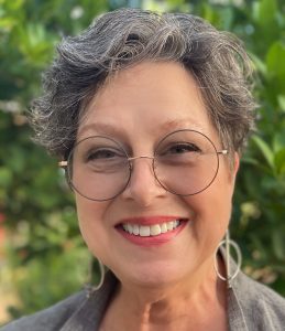 A smiling older woman with short gray hair, round glasses, and large hoop earrings poses outdoors in front of green foliage. She is wearing a gray top and looks directly at the camera.