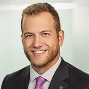 A smiling man with short, light brown hair and a trimmed beard, wearing a dark suit, white shirt, and light purple tie, posed in front of a softly blurred light background.